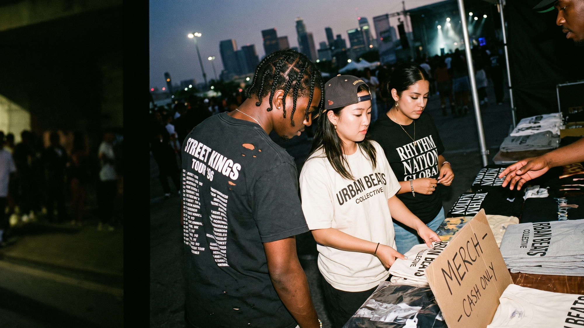 Fans wearing bootleg rap tour tees at a concert merch table at night