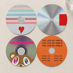 Four Kanye West album-inspired CD-style coasters, full-bleed designs on a light ivory table. One of each design in the set.